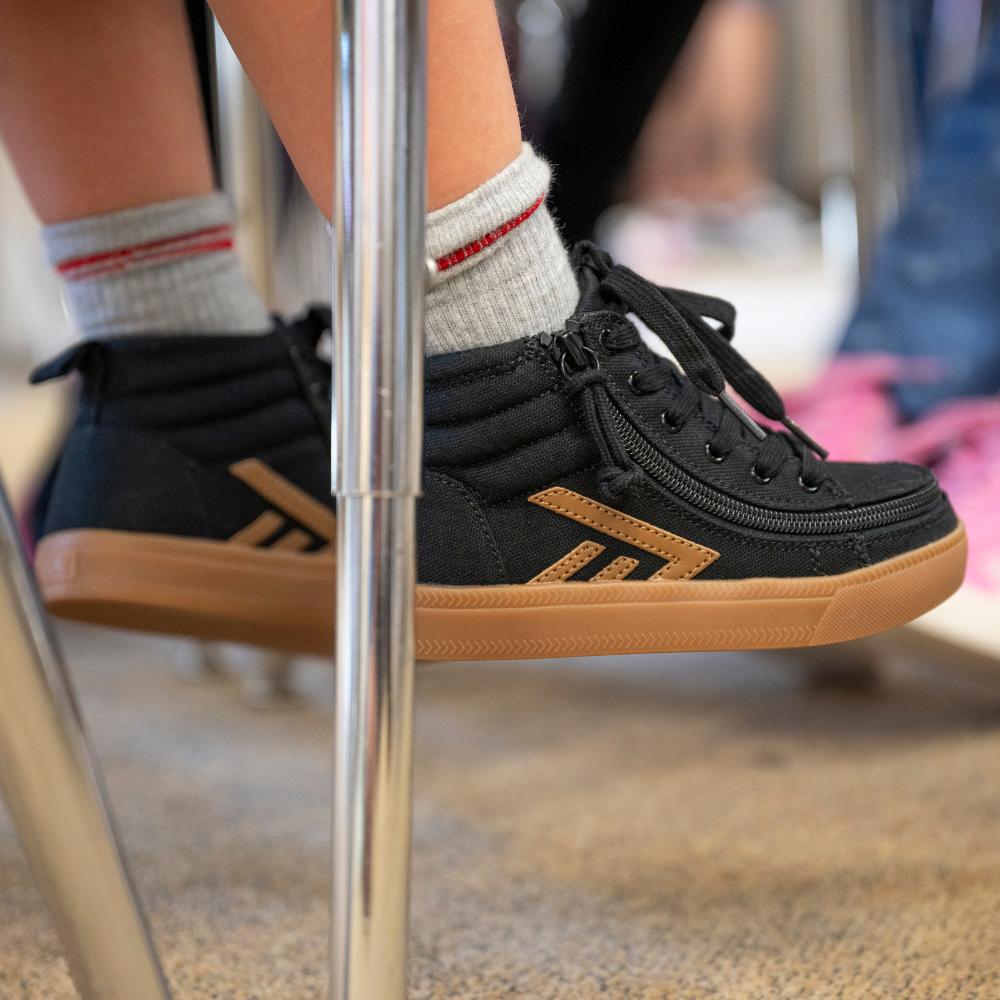 A close-up of a kid wearing black and brown high-top sneakers with grey socks, sitting indoors with feet resting near metal chair legs.