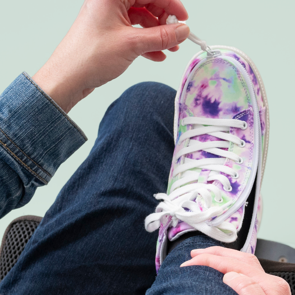 A person wearing jeans and a denim jacket is zipping shoe with white laces on a tie-dye sneaker with purple, green, and white colors.
