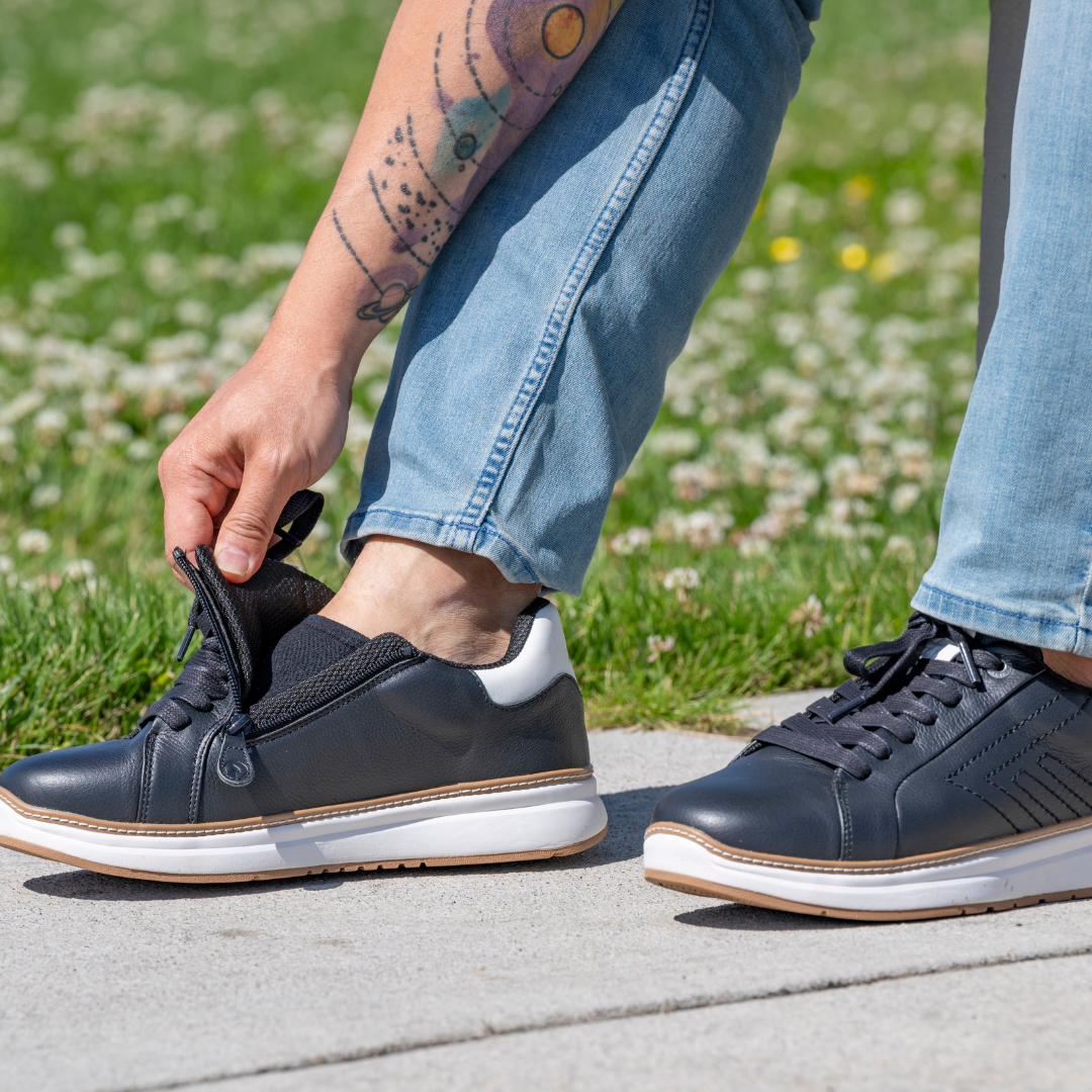 A person in light blue jeans unzips the side of a black sneaker to put it on, revealing a tattooed arm. They are outdoors on a sunny day, standing on a sidewalk next to grass.