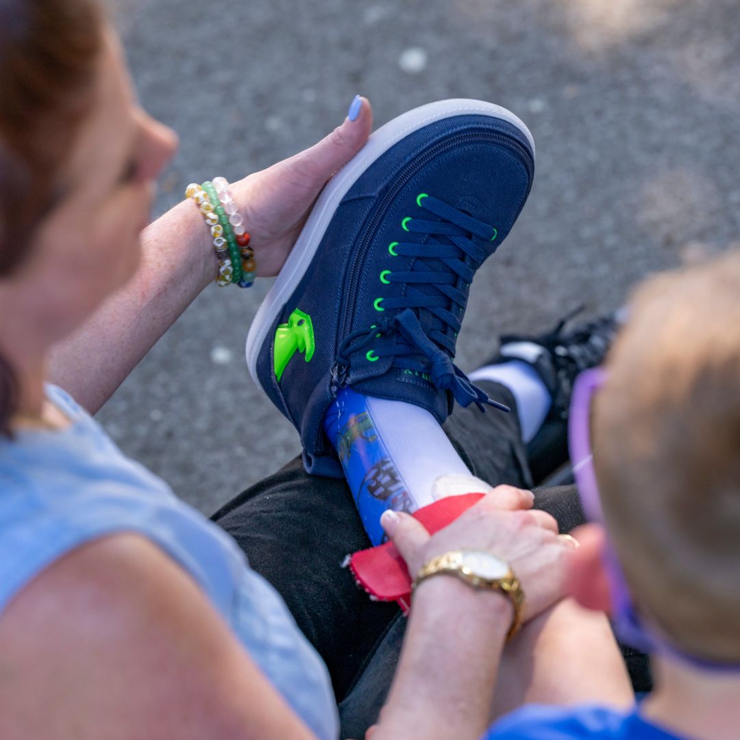 A person helps a child with a orthotic brace put on a blue sneaker with green lace eyelets, sitting outdoors on a sunny day.