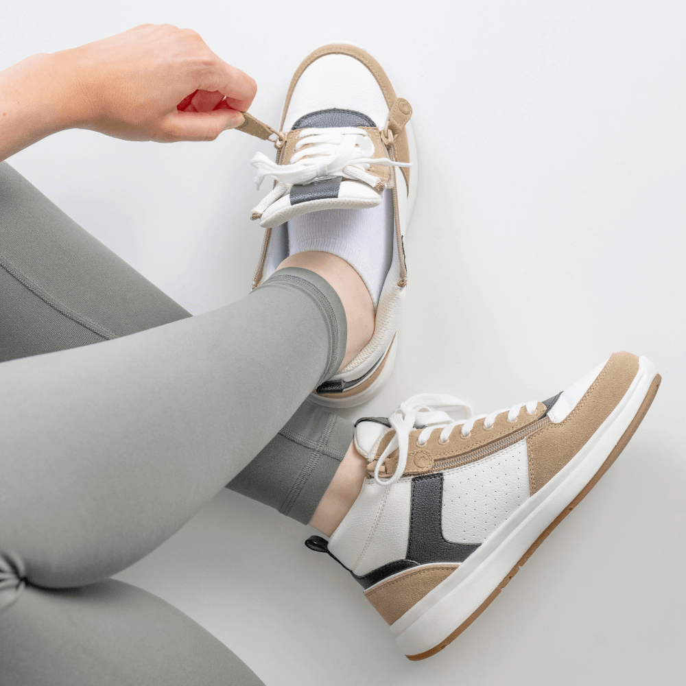 A person wearing grey leggings and white socks ties the laces and zips on beige and white sneakers while sitting on a white surface.