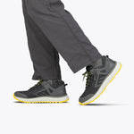 A person wears Men's Charcoal/Yellow BILLY Inclusion Trail sneakers from BILLY Footwear, featuring yellow soles and accents, paired with dark gray pants. Only their lower legs and feet are visible against a light background.