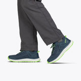 A person in dark gray pants wears BILLY Footwear's Men's Navy/Green BILLY Inclusion Trail—navy athletic slip-on hiking shoes with neon green accents—walking against a light gray background, with only lower legs and feet visible.