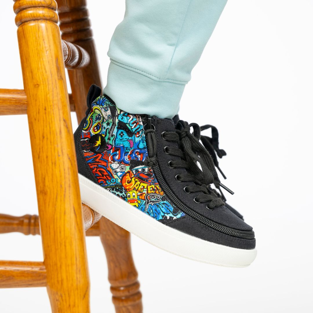 A person in light blue pants stands on a wooden stool, showcasing BILLY Footwear’s Night Sk8ter BILLY Classic D|R II High shoes with graffiti-style artwork on the sides and white soles.