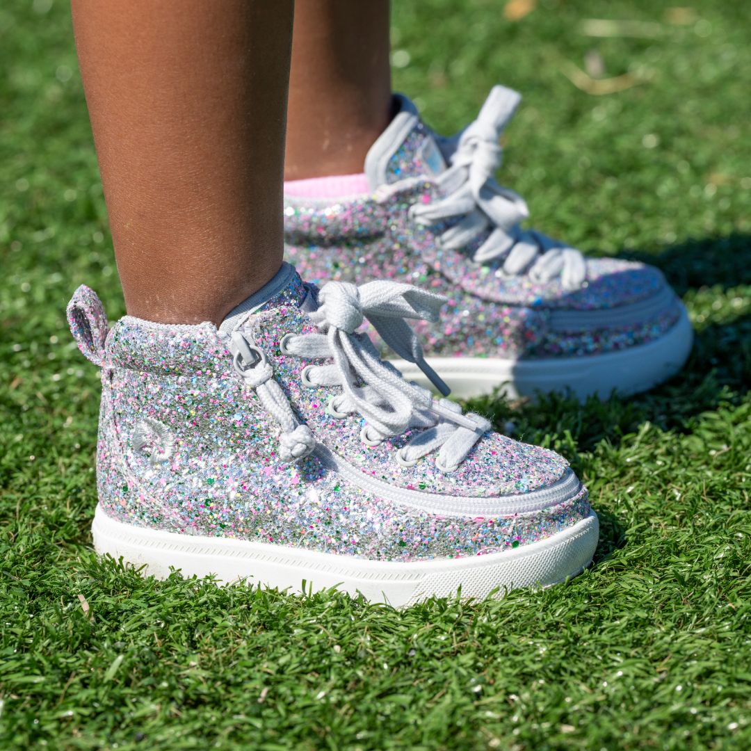 A child wearing BILLY Footwear's Silver Multi Glitter BILLY Classic D|R II High orthotic-friendly sneakers with white laces stands on green artificial grass—perfect kids’ adaptive footwear.